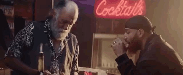 two men are sitting in front of a neon sign that says cocktails