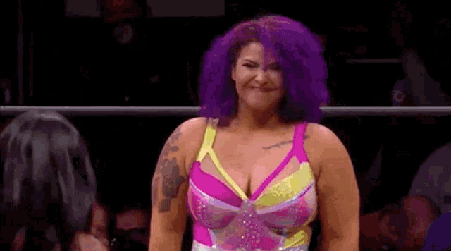 a woman with purple hair is standing in a wrestling ring and smiling at the camera .