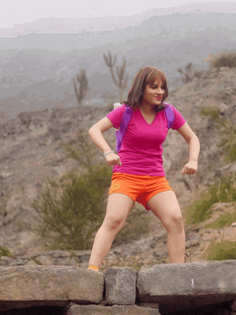 a girl in a pink shirt and orange shorts is jumping on a rock