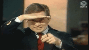 a man in a suit and tie is pointing at the camera while covering his eyes with his hand ..