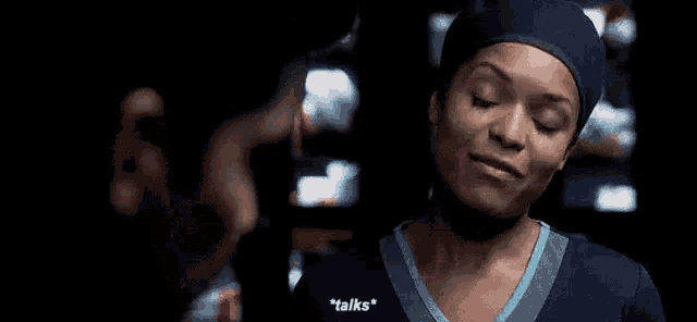 a woman in a scrub top and a hat is talking to a man in a dark room .