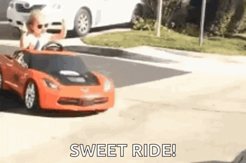 a young boy is driving a red toy car down a street .