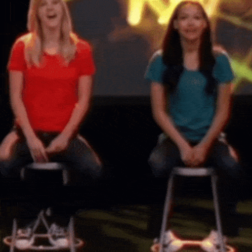 two women are sitting on stools in front of a yellow sign