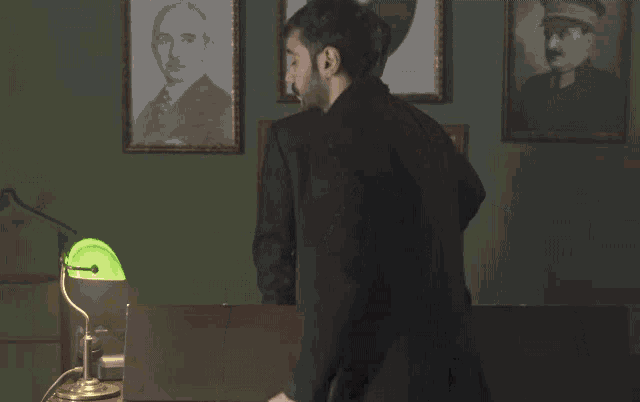 a man with a beard stands in a room with a green lamp