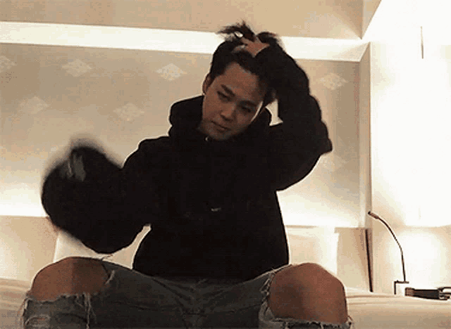 a man in a black hoodie is sitting on a bed adjusting his hair