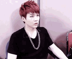 a young man with red hair is sitting in a chair wearing a black shirt and a chain around his neck .