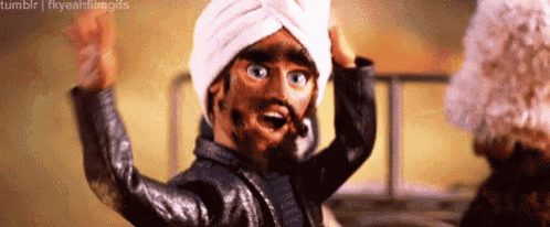 a puppet wearing a turban and a leather jacket is waving his hands .