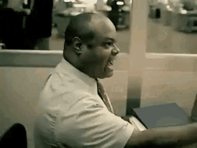 a man in a white shirt and tie is sitting at a desk in an office with his mouth open .