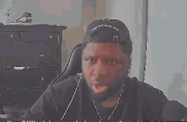 a man wearing a baseball cap is sitting at a desk making a funny face .