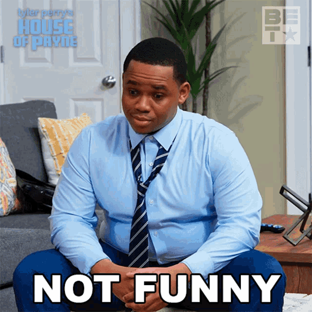 a man in a blue shirt and tie sits on a couch with the words not funny below him