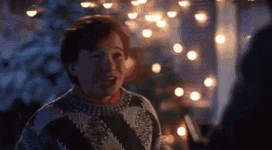 a man in a sweater is standing in front of a christmas tree and talking to another man .
