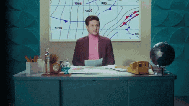 a man in a pink turtleneck is sitting at a desk in front of a weather chart