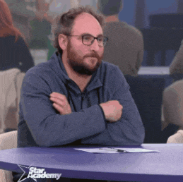 a man with a beard is sitting at a table with his arms crossed in front of a star academy sign