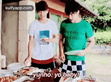 two men are standing next to each other at a table with food and one of them is holding a plate of food .