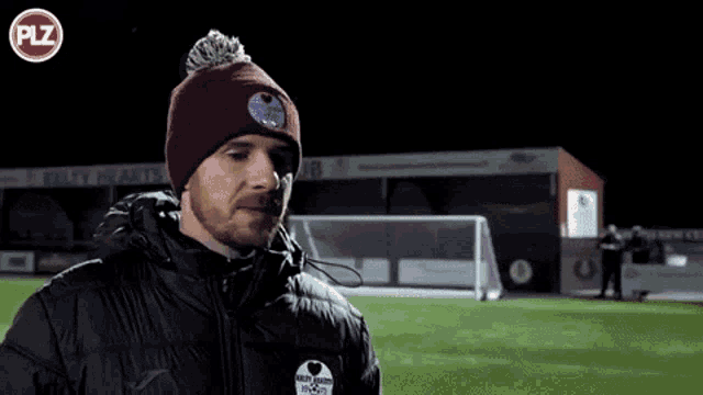 a man with a beard wearing a burgundy beanie with a patch that says ' play hearts foot '