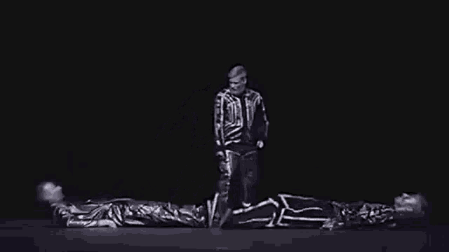a black and white photo of a man in a skeleton costume standing next to two people laying on the floor .