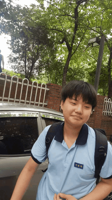 a boy wearing a blue shirt with a black collar and a black backpack stands in front of a car