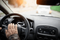 a person is driving a car on a city street and their hand is on the steering wheel .
