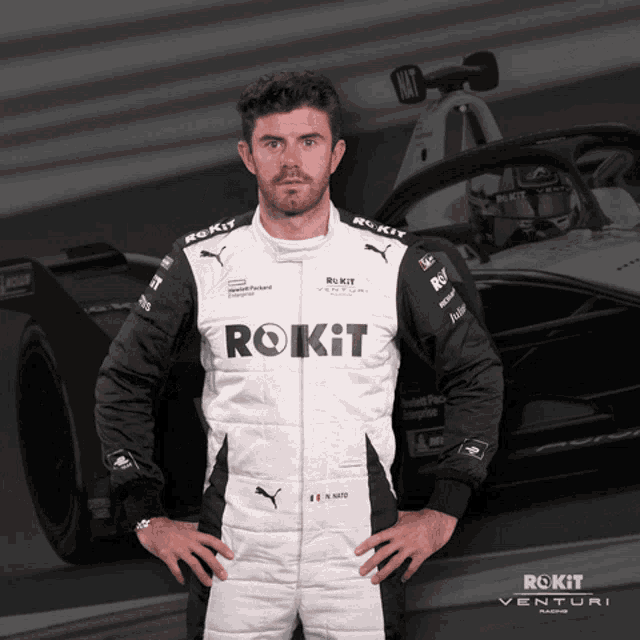 a man wearing a rokit racing suit stands in front of a race car
