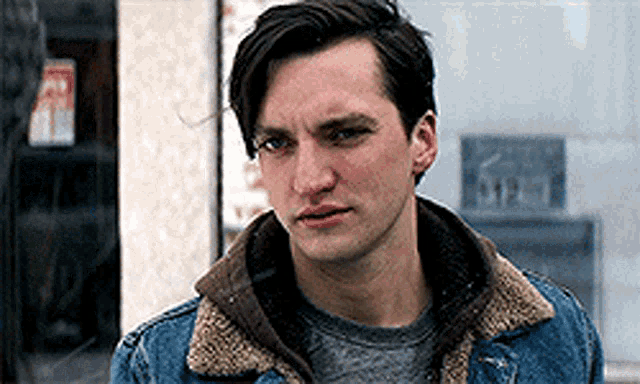 a young man wearing a denim jacket and a sweater is standing in front of a building .
