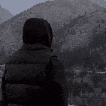 a person in a hooded jacket stands in front of a mountain range
