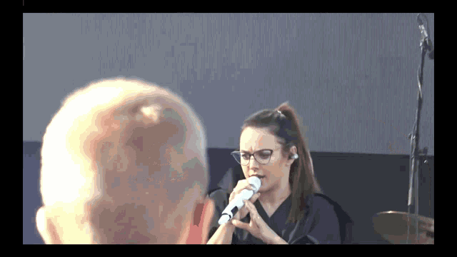 a woman singing into a microphone with a bald head in the background