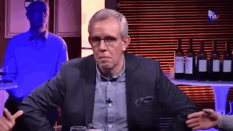 a man in a suit and glasses is sitting at a table with bottles of wine on it and a tv logo in the corner