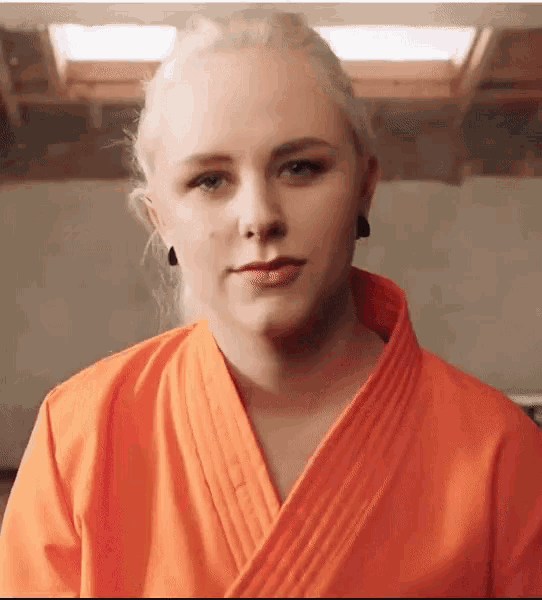 a blonde woman in an orange karate uniform looks at the camera