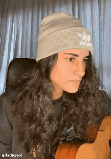 a woman with long hair wearing an adidas beanie holds a guitar