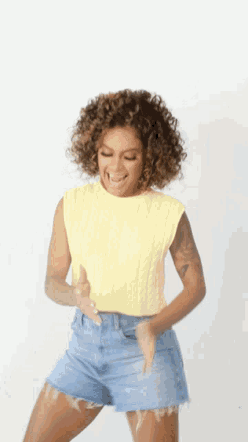 a woman with curly hair wearing a yellow top and blue shorts is dancing