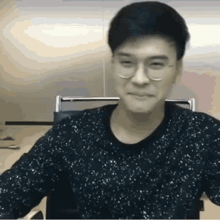 a man wearing glasses and a black sweater is sitting in a chair and smiling .