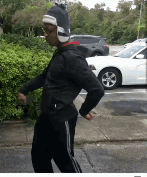 a man wearing a black jacket and a hat is dancing in front of a white car in a parking lot