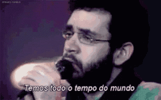 a man with glasses is singing into a microphone with the words temos todo o tempo do mundo above him .