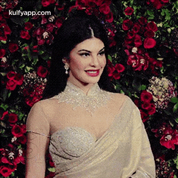 a woman in a white saree is smiling in front of a wall of flowers .
