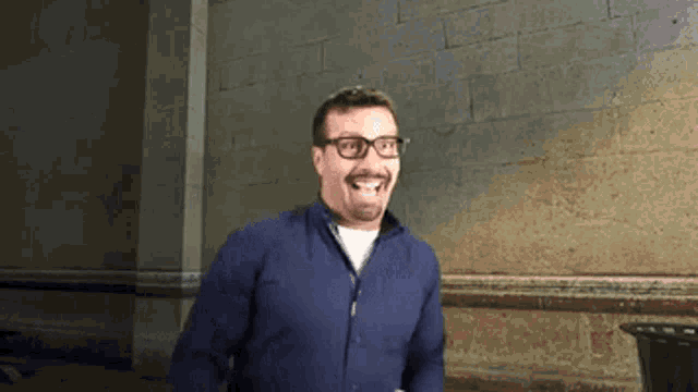 a man wearing glasses and a blue shirt stands in front of a brick wall