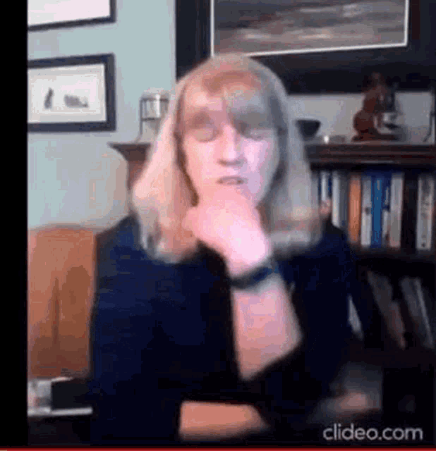 a woman is sitting in front of a bookshelf with her eyes closed and her hand on her chin .