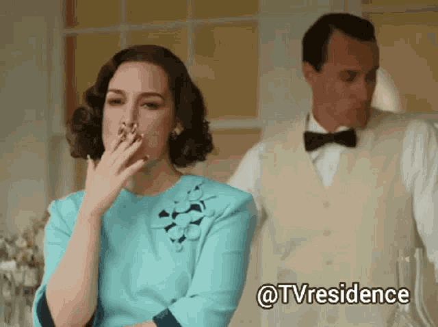 a woman in a blue dress is covering her mouth with her hand while a man in a white vest stands behind her