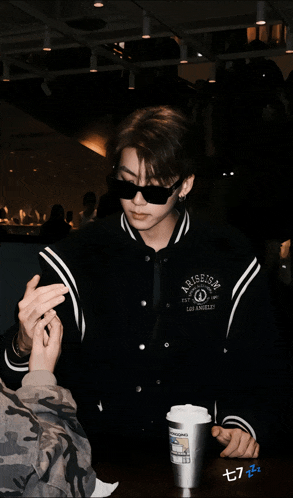 a man wearing sunglasses and a jacket that says arise on it sits at a table with a cup of coffee