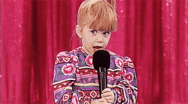 a little girl in a floral dress is holding a microphone in front of a pink curtain .