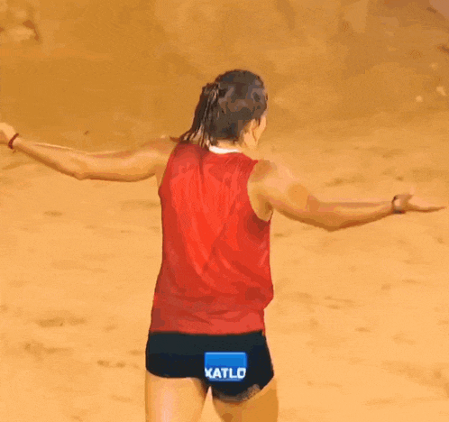 a woman in a red tank top and black shorts is standing on a sandy beach with a sign that says exatlon mexico