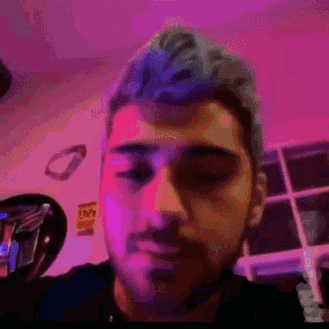 a man with blue hair and a beard looks at the camera in a room with purple lights