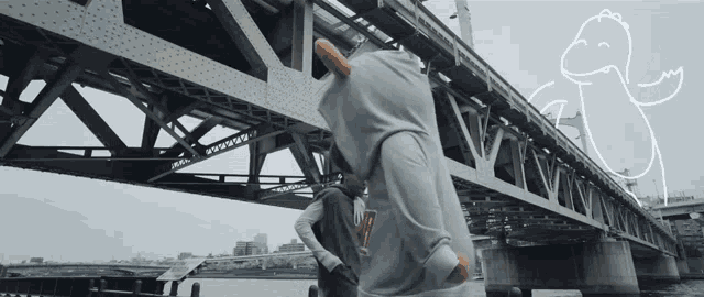 a person wearing a dinosaur hoodie stands in front of a bridge with a drawing of a dinosaur behind them