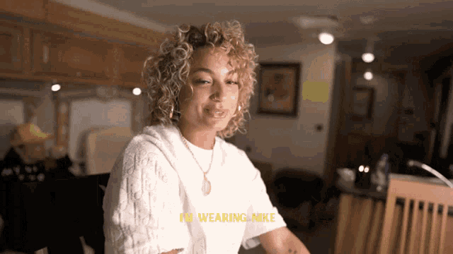 a woman with curly hair is wearing a white sweater and a gold necklace and says i 'm wearing nike