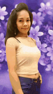 a woman in a white tank top stands in front of a purple background of flowers