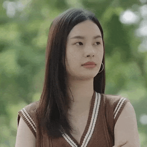 a woman wearing a brown vest and hoop earrings is standing in a park with her arms crossed .