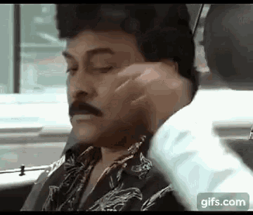 a man with a mustache is sitting in a car with his eyes closed and a hand on his face .