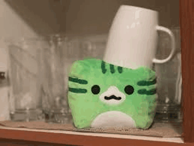 a green cat stuffed animal is sitting on a shelf next to a white cup .
