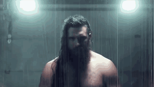 a shirtless bearded man is standing in the rain in a dark room .