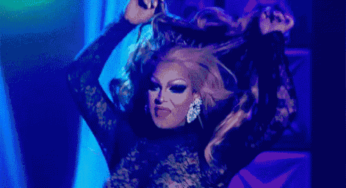 a drag queen is standing on a stage with her arms outstretched and making a funny face .
