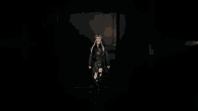 a woman is walking down a runway in a dark room wearing a black leather dress and boots .